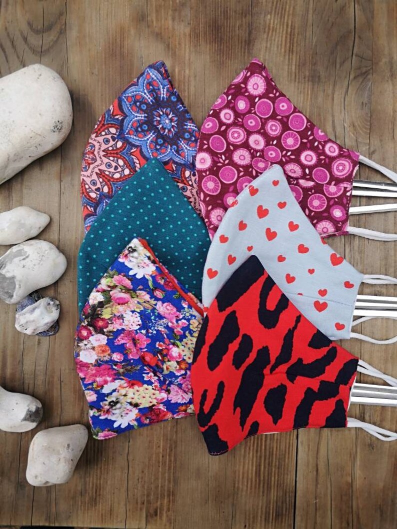 May include: Six different fabric face masks with various patterns. The masks are made of cotton and have a variety of colors, including red, blue, green, pink, and white. The masks are all folded and arranged in a pile on a wooden surface.
