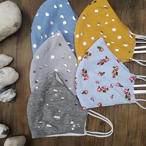 May include: Five fabric face masks with different patterns. The masks are all light blue, gray, or yellow with silver polka dots or a floral pattern. The masks are all made of a soft, breathable fabric.