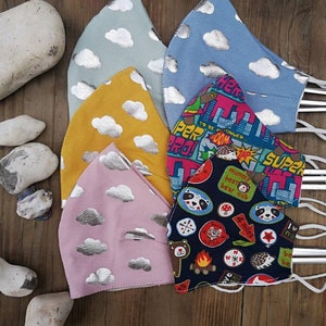 May include: Five fabric face masks with different patterns. The masks are all made of a soft, cotton fabric and have adjustable ear loops. The patterns include clouds with silver accents, a cityscape with the text "To Be Continued..." and "Super Pro!" and a camping theme with a variety of colorful patches.