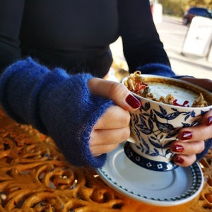 May include: A pair of fuzzy, deep blue fingerless gloves made of a soft, textured knit material. The gloves cover the wrists and the back of the hands, leaving the fingers exposed. The knit has a visible, slightly brushed, and airy texture.