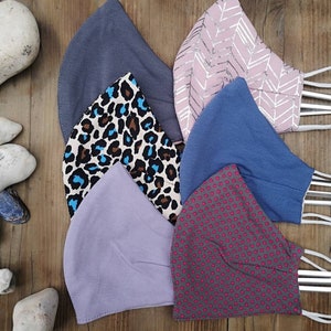 May include: Six different fabric face masks. The masks are all made of cotton and have different patterns. The patterns include a gray solid, a pink chevron, a blue solid, a brown and blue leopard print, a purple solid, and a gray with pink polka dots.