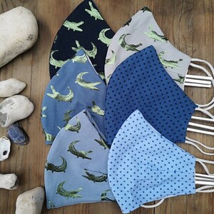 May include: Five different fabric face masks with various patterns. The masks are blue, gray, and beige with green alligator designs and blue polka dots. The masks are made of cotton and have elastic ear loops.