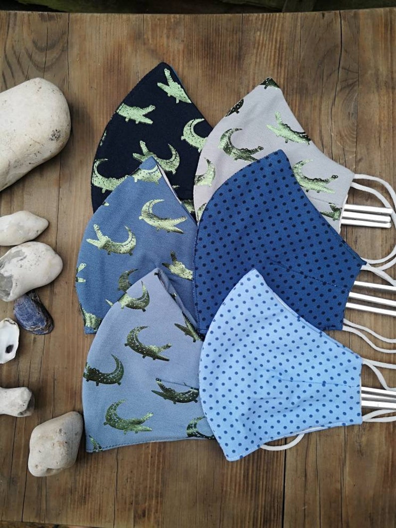 May include: Five different fabric face masks with various patterns. The masks are blue, gray, and beige with green alligator designs and blue polka dots. The masks are made of cotton and have elastic ear loops.