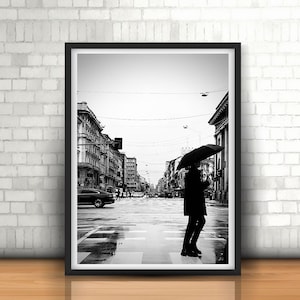 以下が含まれることがあります： 傘を持った人が街の通りを歩いている白黒写真。その人はコートを着ており、雨の中を歩いている。通りは濡れていて、通りの両側に建物がある。