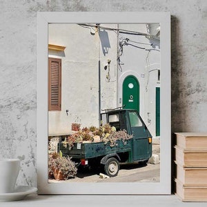 May include: A vintage green truck with a wooden bed is parked in a narrow alleyway. The truck is filled with potted plants and flowers. The alleyway is lined with white buildings with a green door and a brown window.