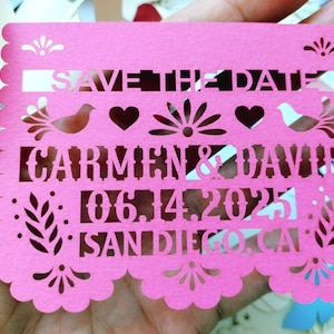 May include: A pink paper cutout with scalloped edges, featuring the text "SAVE THE DATE", names "CARMEN & DAVID", and the date "06.14.2025" along with "SAN DIEGO, CA". The design includes decorative birds, hearts, and floral elements.