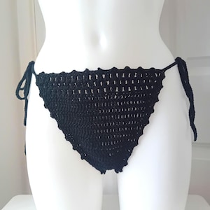 May include: Black crochet bikini bottoms with tie sides. The bottoms are made of a dark, solid black yarn and feature a scalloped edge along the top. The ties are long and can be adjusted for a comfortable fit. The bottoms are displayed on a white mannequin.