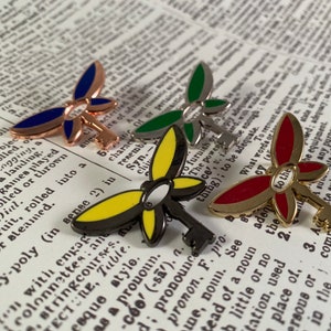 May include: Four enamel pins featuring a key with dragonfly wings. The wings are in different colors: blue, green, yellow, and red.
