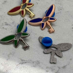 K&ouml;nnte beinhalten: Vier metallene Schmetterlings-Schl&uuml;sselnadeln mit unterschiedlich farbigen Fl&uuml;geln. Die Fl&uuml;gel sind rot, blau, gr&uuml;n und silber. Die Schl&uuml;ssel sind alle silber.