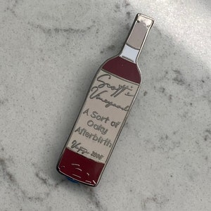 May include: A silver and red enamel pin shaped like a wine bottle. The label reads "Scoff's Vineyard, A Sort of Oaky Afterbirth, V+77 2005".