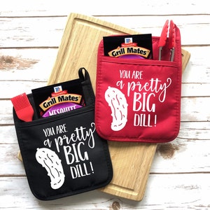 May include: Two pot holders, one black and one red, with the text "YOU ARE a pretty BIG DILL!" and a pickle graphic. Each holds a packet of Grill Mates seasoning and a grilling utensil. A wooden cutting board is in the background.