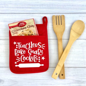 May include: Red oven mitt with a pocket that holds a package of chocolate chip cookie mix. The oven mitt has the text "Teachers Bake Smart Cookies" with a rolling pin graphic. Two wooden kitchen utensils are on a white wood background.