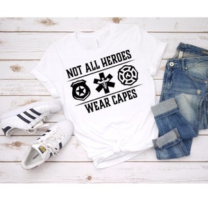 May include: White t-shirt with black text that reads "Not all heroes wear capes". The text is surrounded by three symbols: a police badge, a medical symbol, and a fire department symbol. The shirt is laid out on a wooden surface with a pair of white sneakers and blue jeans.