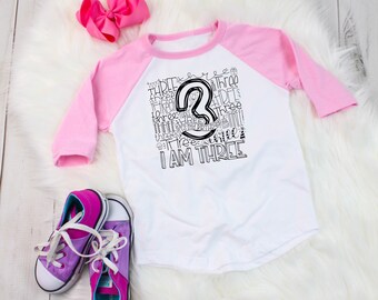 May include: A white and pink baseball tee shirt with a black and white graphic design that says "I am three" with the number 3 in the center. The shirt is on a white fuzzy surface with a pink bow and purple sneakers.