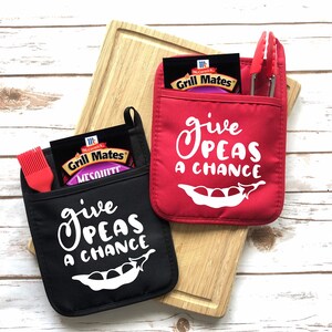 May include: Two pot holders, one black and one red, with the phrase "Give Peas a Chance" in white script. The black pot holder contains a red silicone brush and a packet of Grill Mates Mesquite seasoning. The red pot holder contains red tongs.