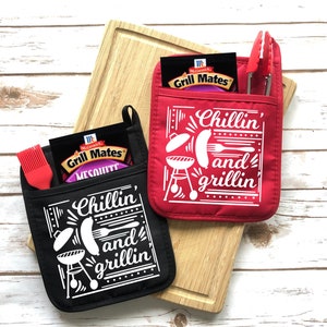 May include: Two red and black oven mitts with a pocket on the front. The mitts have a white design that says "Chillin' and grillin'" with a grill, hot dog, and other grilling icons.