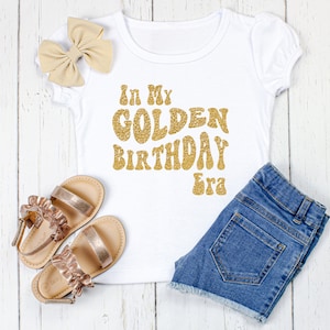 May include: White t-shirt with gold glitter lettering that says "In My Golden Birthday Era".