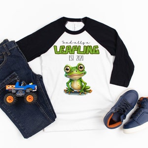 May include: A white and black baseball tee shirt with a green frog graphic and the text "Toad-ally a Leapling Est. 2020".