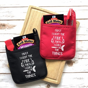 May include: Two red and black oven mitts with pockets. The mitts have a white graphic that says "First I light the fire then I grill the things".