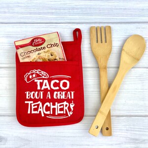 May include: Red oven mitt with a pocket that holds a package of chocolate chip cookie mix. The oven mitt has white text that says "Taco 'bout a great teacher!" and a taco graphic. Two wooden kitchen utensils are in the background.