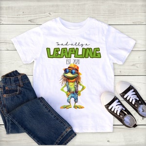 May include: White t-shirt with a green frog wearing sunglasses and a hat. The shirt has the text "Toad-ally a Leapling EST 2020" printed on it.