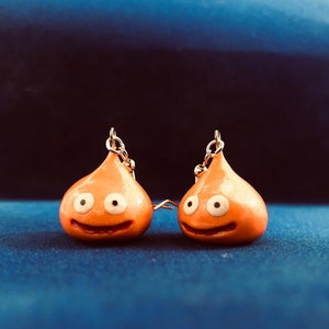 May include: A pair of orange slime earrings with white eyes and a smiling mouth. The earrings are hanging from silver hooks.