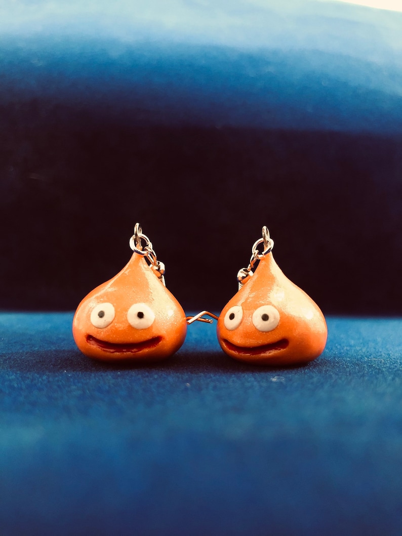 May include: A pair of orange slime earrings with white eyes and a smiling mouth. The earrings are hanging from silver hooks.