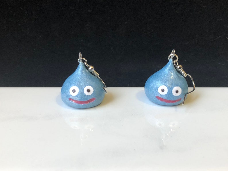 May include: A pair of blue and silver earrings featuring a smiling slime character with white eyes.