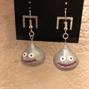 May include: A pair of silver dangle earrings featuring a slime character with a smiling face and white eyes. The earrings are on a black card.
