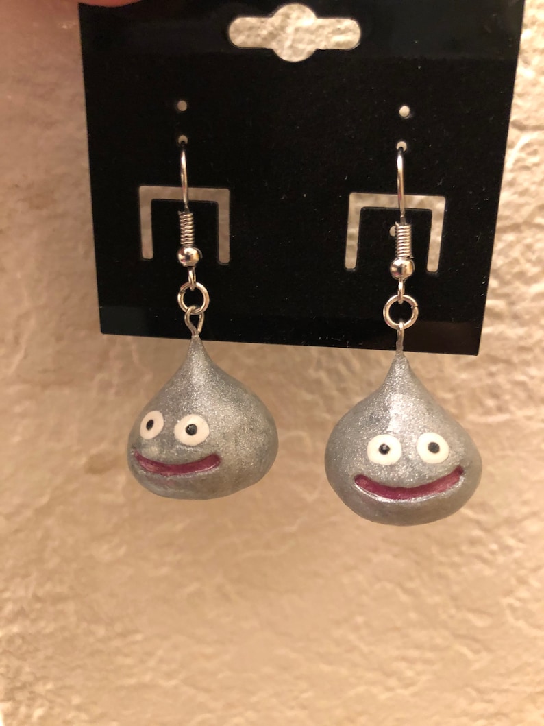 May include: A pair of silver dangle earrings featuring a slime character with a smiling face and white eyes. The earrings are on a black card.