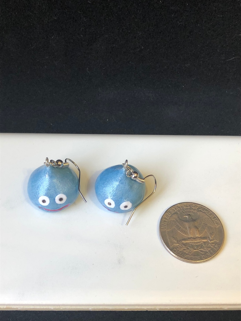 May include: A pair of blue slime earrings with white eyes and a red mouth. The earrings are about the size of a US quarter.