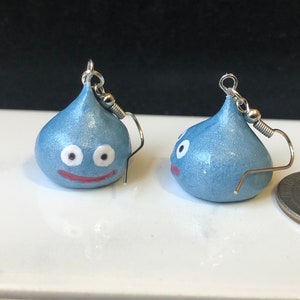 May include: A pair of blue slime earrings with white eyes and a red mouth. The earrings are made of polymer clay and are about the size of a US quarter.