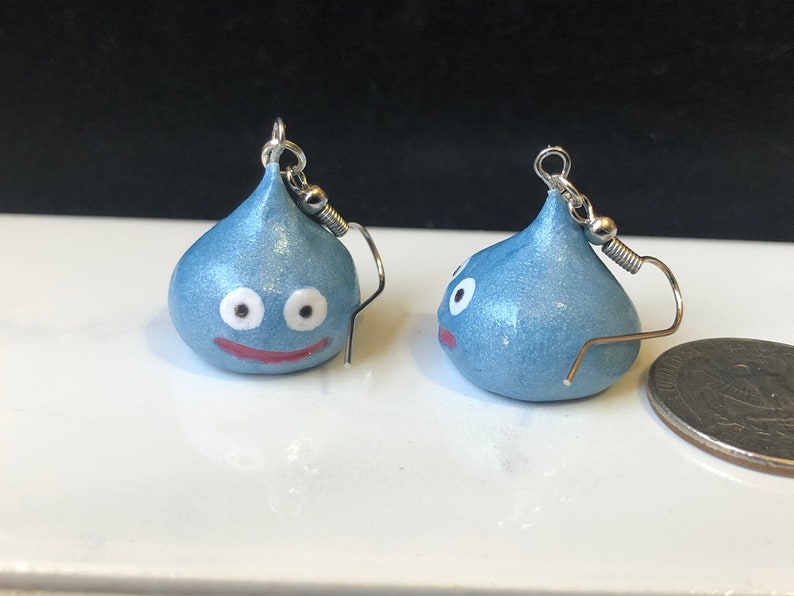 May include: A pair of blue slime earrings with white eyes and a red mouth. The earrings are made of polymer clay and are about the size of a US quarter.