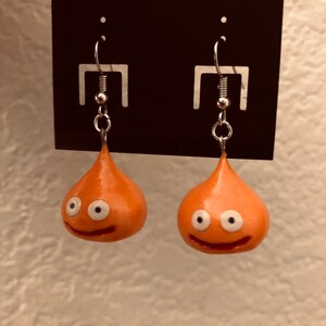 May include: A pair of orange slime earrings with white eyes and a smiling mouth. The earrings are on a black card with a silver hook.