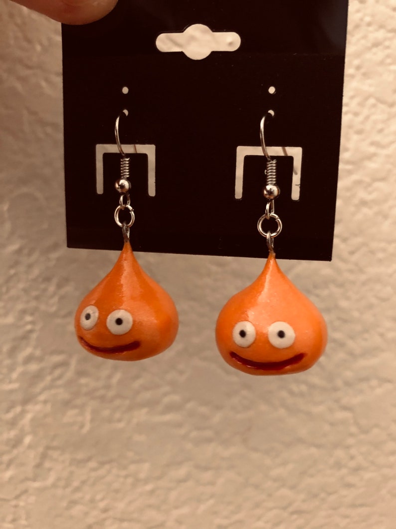 May include: A pair of orange slime earrings with white eyes and a smiling mouth. The earrings are on a black card with a silver hook.