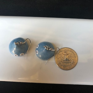 May include: A pair of blue ceramic earrings with white dots, resembling eyes, hanging from silver hooks. A US quarter is included for size reference.