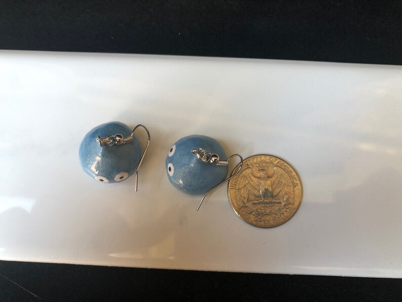 May include: A pair of blue ceramic earrings with white dots, resembling eyes, hanging from silver hooks. A US quarter is included for size reference.