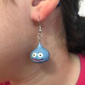 May include: A pair of silver dangle earrings with a blue and white slime character charm. The charm is shaped like a teardrop with a smiling face and two white eyes.