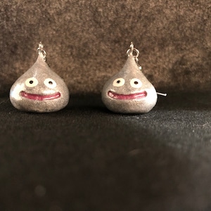 May include: A pair of silver earrings shaped like a smiling slime monster with white eyes and a pink mouth.