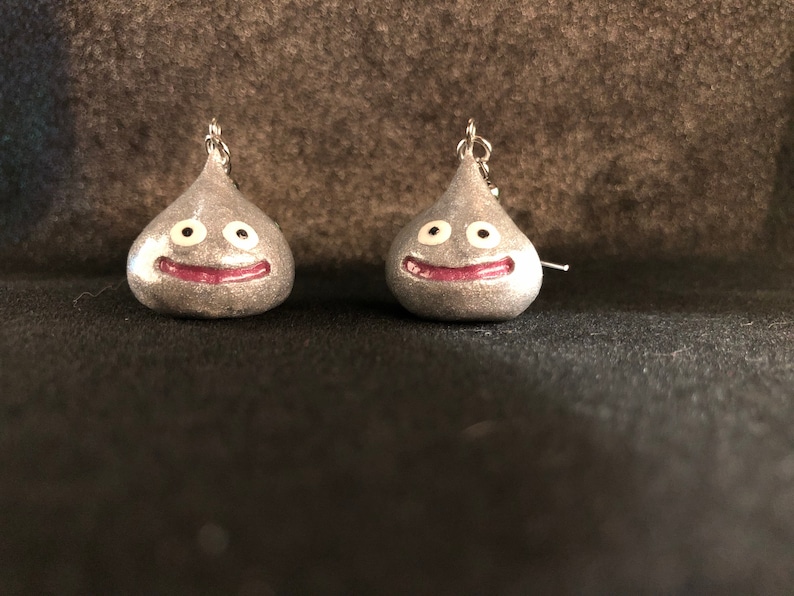 May include: A pair of silver earrings shaped like a smiling slime monster with white eyes and a pink mouth.