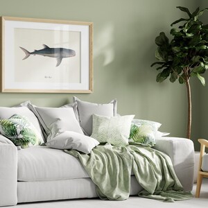 Pu&ograve; includere: Un divano bianco con cuscini e una coperta verde. Il divano &egrave; di fronte a un muro verde salvia con una stampa incorniciata di uno squalo. Una pianta verde frondosa si trova nell'angolo della stanza.