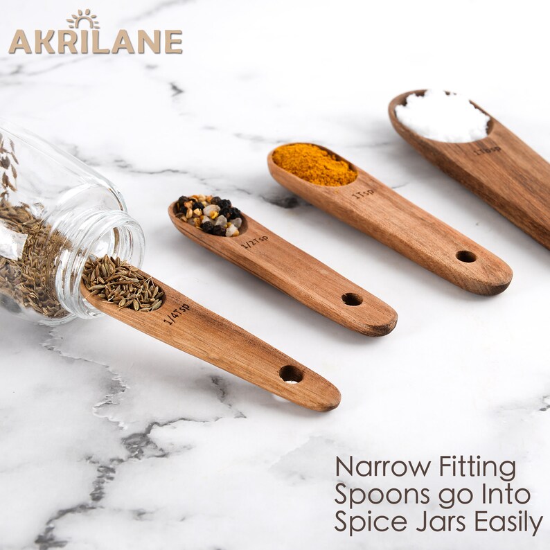May include: Four wooden spoons with measurements marked on them. The spoons are filled with different spices, including cumin, turmeric, and salt. The spoons are on a white marble surface. The text "Narrow Fitting Spoons go Into Spice Jars Easily" is printed below the spoons.