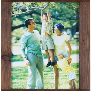 May include: A rectangular picture frame made of dark brown stained wood with visible grain. The frame features four corners accented with light-colored twine tied through small holes, enclosing a photograph of a family in a park.