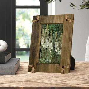 May include: A rectangular picture frame made of weathered brown wood with a visible grain. The frame features thick borders and is accented with natural jute rope tied around each of the four corners. It contains a photograph of white flowers.