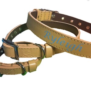 Op de afbeelding: Lichtbruine leren hondenhalsbanden met zilverkleurige gespen en D-ringen. Eén halsband is gepersonaliseerd met de naam "Ryleigh" in lichtblauwe letters. De halsbanden hebben een bruine binnenkant en een klassieke gespsluiting.