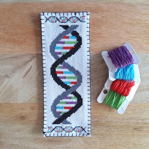 May include: A white cross-stitch bookmark with a DNA double helix design in black, gray, red, purple, blue, and green. The bookmark is surrounded by a black border. Four spools of embroidery floss are shown in the background, in purple, blue, red, and green.
