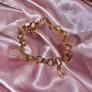 May include: A gold chain anklet with a pink butterfly charm. The anklet is on a pink satin fabric background.
