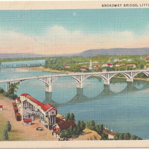 May include: Vintage postcard featuring the Broadway Bridge in Little Rock, Arkansas. The bridge is white with arched supports, spanning a wide river. The postcard has a blue sky and a town in the background.