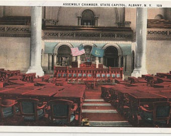 May include: Vintage postcard of the Assembly Chamber in the State Capitol, Albany, NY. The image shows rows of red desks and chairs, with a raised platform at the front. Two flags are displayed above the platform. The text "ASSEMBLY CHAMBER. STATE CAPITOL, ALBANY, N. Y. 109" is at the top.