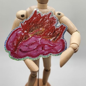 May include: A wooden artist's mannequin holds a red and pink abstract shape with glitter accents and a holographic border. The artwork is a vibrant mix of colors and textures, creating a unique visual piece.